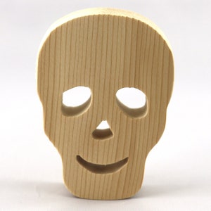 A handcrafted, unfinished light wood Halloween skull cutout featuring carefully carved eye sockets, a nose, and a cheerful, smiling mouth. Displayed upright against a white background.