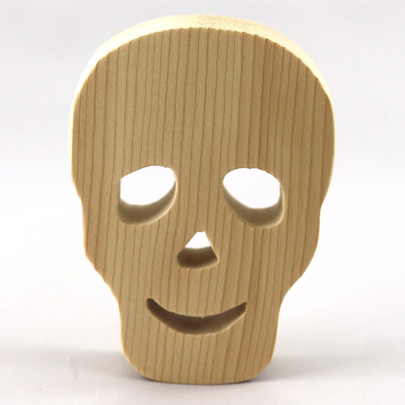 A handcrafted, unfinished light wood Halloween skull cutout featuring carefully carved eye sockets, a nose, and a cheerful, smiling mouth. Displayed upright against a white background.
