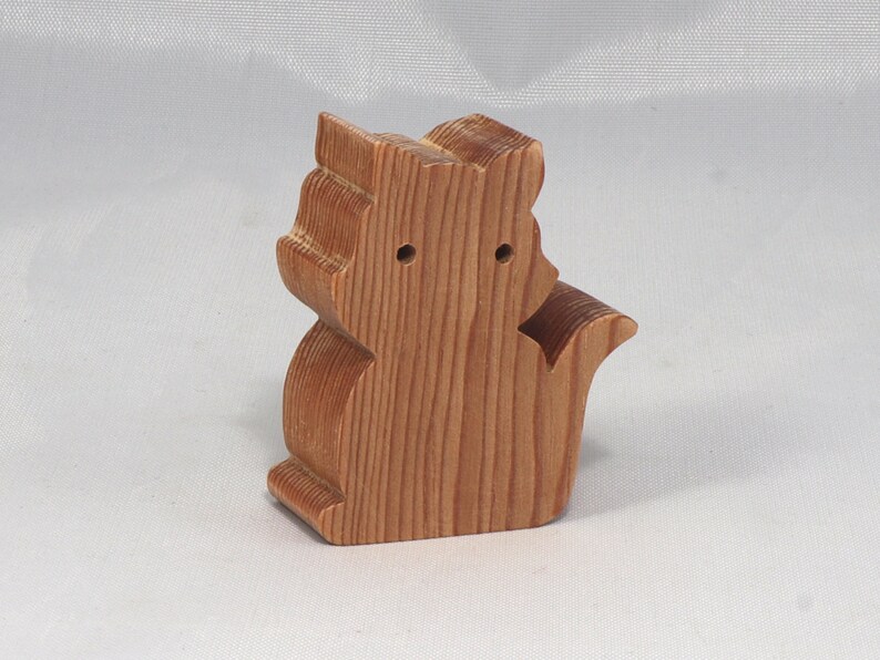 May include: A wooden toy squirrel figurine with a light brown finish. The squirrel is standing on its hind legs with its tail curled behind it.