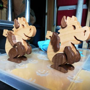 Two wooden baby dragons on a workshop table, both facing right, with light wood bodies and contrasting dark wood wings and legs.