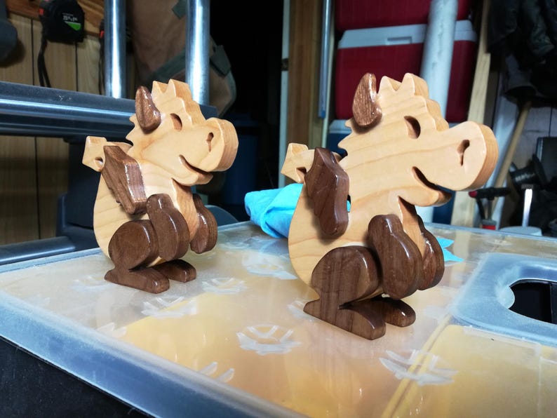 Two wooden baby dragons on a workshop table, both facing right, with light wood bodies and contrasting dark wood wings and legs.