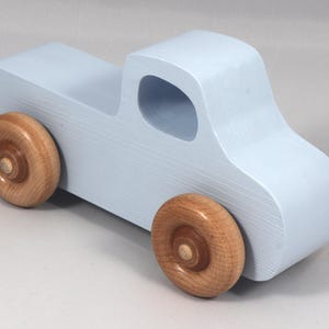 May include: A light blue wooden toy truck with natural wood wheels. The truck has a classic pickup design with a cab and a cargo bed. The wheels are round and attached to the truck's body.
