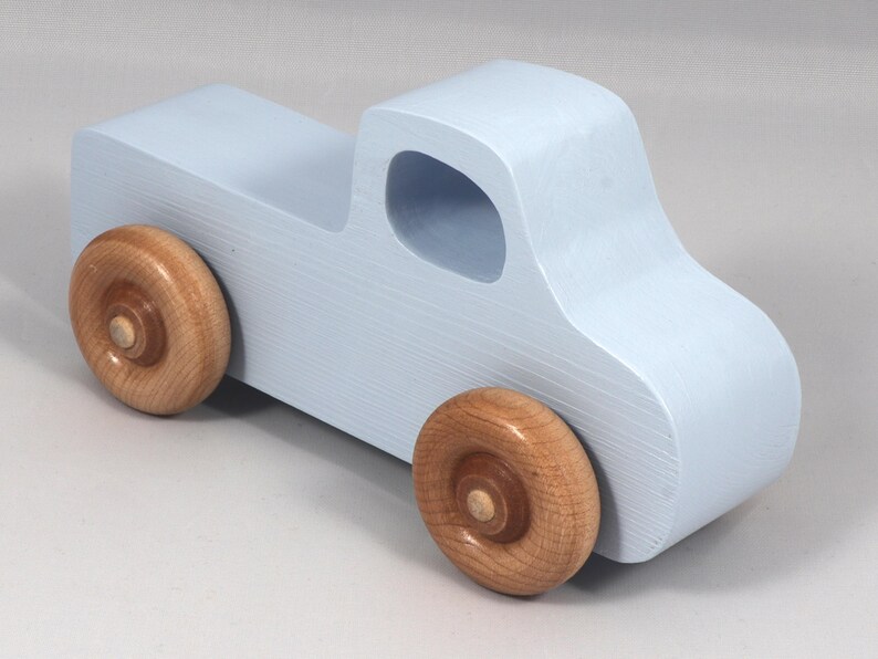 May include: A light blue wooden toy truck with natural wood wheels. The truck has a classic pickup design with a cab and a cargo bed. The wheels are round and attached to the truck's body.
