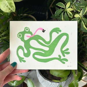 May include: Green frog with a pink tongue catching a black fly. The frog is depicted in a stylised, blocky design. The image is a print on white paper.