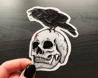 May include: A black crow perched on top of a white human skull with a red blood splatter on the forehead.