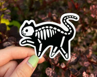 May include: A black and white sticker featuring a cat skeleton design. The sticker has a white border and is held against a backdrop of green and burgundy foliage. The cat skeleton design includes the skull, ribs, spine, and legs.