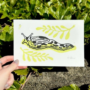 May include: A black and white illustration of a slug on a white background with yellow leafy branches. The slug is outlined in yellow and has black spots. The artist's signature is in the bottom right corner.