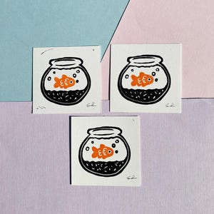 May include: Three black and white illustrations of a goldfish in a bowl. The goldfish is orange and has a black eye. The bowl is filled with black and white dots to represent water.