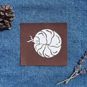 Peut inclure: Un écusson carré marron avec une illustration blanche d'un cloporte. Le dessin du cloporte est centré sur l'écusson. L'écusson est sur une surface en denim bleu, avec une pomme de pin et une brindille de lavande en arrière-plan.