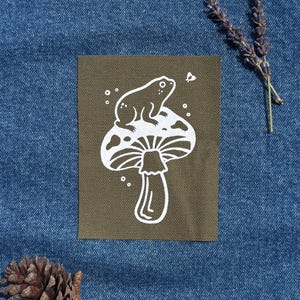 May include: An olive green patch with a white illustration of a frog on a mushroom. The patch is rectangular and set against a blue denim background. A small butterfly and dots are also included in the design.