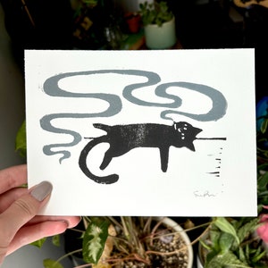 May include: Black and white print of a cat with a grey smoke design behind it. The print is signed "Suhni".