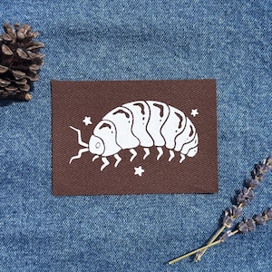 Peut inclure: Un écusson marron avec une illustration blanche d'un cloporte, montrant son corps segmenté et ses antennes. Le motif comprend de petits accents d'étoiles. L'écusson est posé sur un fond en denim bleu, avec une pomme de pin et une brindille de lavande.