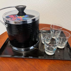 May include: A black ice bucket with a silver rim and handle, set on a black tray with four shot glasses. The ice bucket has a textured exterior and a colorful design on the lid.