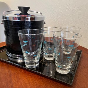 May include: A vintage bar set featuring a black ice bucket with a silver handle and rim, and four clear glasses decorated with blue fish designs. The set is displayed on a black rectangular tray, perfect for serving drinks.
