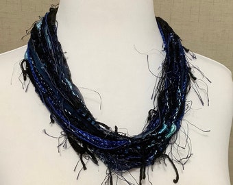 May include: A blue and black yarn necklace with a frayed, textured look. The necklace is made of multiple strands of yarn that are twisted and knotted together.