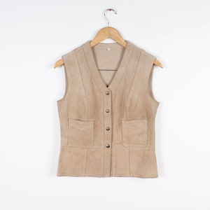 Può includere: Un gilet in pelle scamosciata beige con scollo a V, chiusura con bottoni e due tasche frontali. Il capo senza maniche è appeso a una gruccia di legno su sfondo bianco. Il capo ha un design classico e su misura.