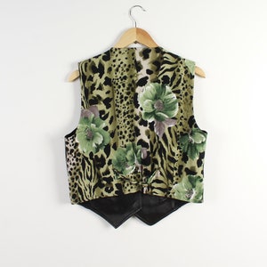 May include: A vintage vest with a distinctive design. The vest showcases a leopard print pattern in green and black, complemented by floral accents. It's displayed on a wooden hanger against a white backdrop.