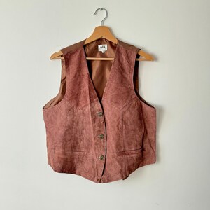 May include: A vintage, dusty rose suede vest with a V-neck and button closure. The vest has a brown satin lining and is hanging on a wooden hanger. The vest has two front pockets and a brown leather yoke.