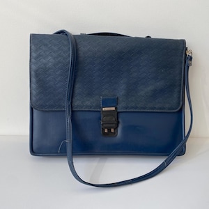 Op de afbeelding: Een marineblauwe leren aktetas met een geweven bovenklep en een schouderriem. De tas heeft een rechthoekige vorm met een metalen sluiting. De tas staat op een wit oppervlak.