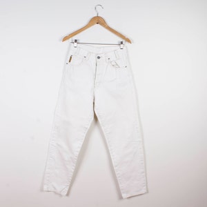 May include: A pair of high-waisted white denim jeans featuring a button fly closure, belt loops, and a small rectangular gold-toned brand tag on the front right pocket area. The jeans are displayed hanging on a wooden hanger against a plain background.