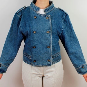 May include: A blue denim jacket with a mandarin collar and double breasted closure. The jacket has four gold buttons and a cropped length.