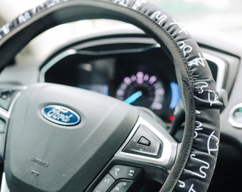 Peut inclure: Un couvre-volant noir avec du texte blanc et un motif de petites formes blanches. Le couvre-volant est sur un volant en cuir noir avec un logo Ford et des boutons.