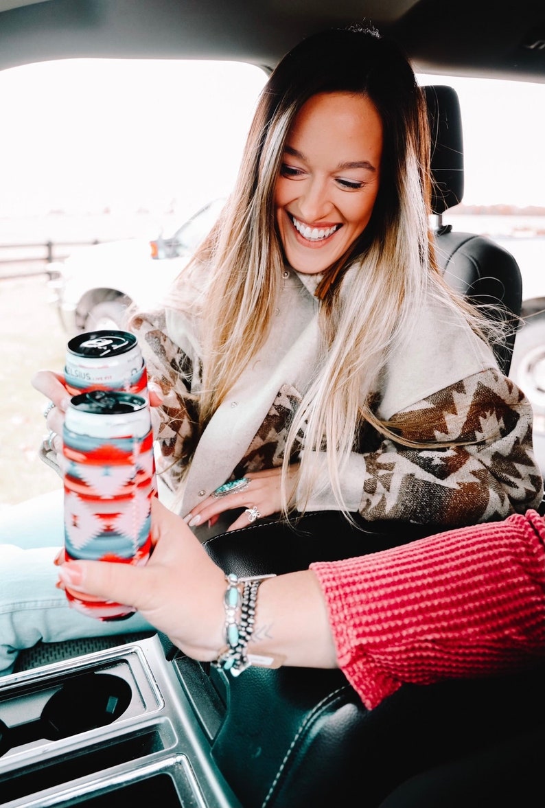 May include: Two aluminum beverage cans with colorful patterned sleeves. The cans are being held by a person's hand in a car.
