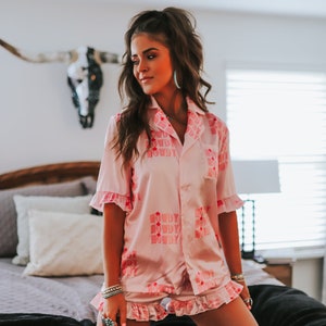 K&ouml;nnte beinhalten: Eine Frau tr&auml;gt einen rosa Satin-Pyjama mit einem "Howdy"-Druck. Der Pyjama hat kurze &Auml;rmel und einen R&uuml;schen-Saum. Die Frau kniet auf einem Bett mit einer schwarzen Decke.