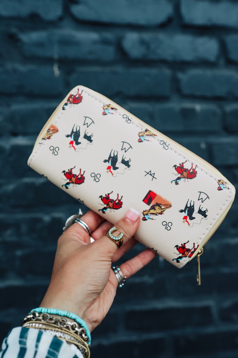 May include: A beige wallet with a zipper closure features a repeating pattern of cowboys riding horses. The pattern includes the letters "98" and "W" and a symbol that looks like a cross with a line through it.