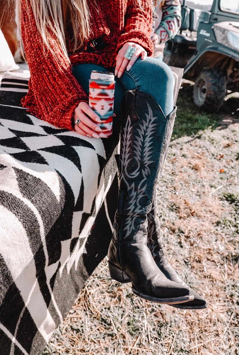 May include: A woman wearing a red sweater and blue jeans sits on a white and black blanket. She is holding a black and white patterned cup with a red and white design. She is wearing black cowboy boots with a floral design.