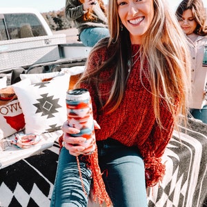 May include: A woman wearing a red sweater and blue jeans is holding a can cooler with a colorful geometric pattern. She is sitting on a black and white patterned blanket with pillows behind her.