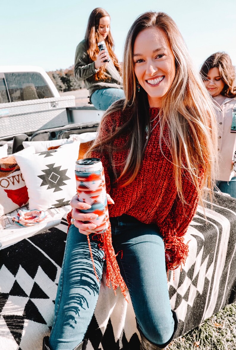 May include: A woman wearing a red sweater and blue jeans is holding a can cooler with a colorful geometric pattern. She is sitting on a black and white patterned blanket with pillows behind her.