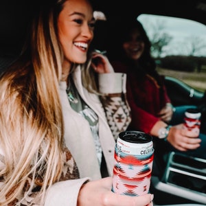 May include: Two women in a car, one holding a red, pink, and blue patterned can cooler with the text "Celsius" on it.