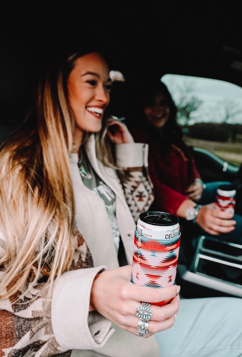 May include: Two women in a car, one holding a red, pink, and blue patterned can cooler with the text "Celsius" on it.