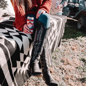 May include: A woman wearing a red sweater and blue jeans sits on a black and white patterned blanket. She is holding a can with a colorful patterned sleeve. She is wearing black cowboy boots with intricate designs.