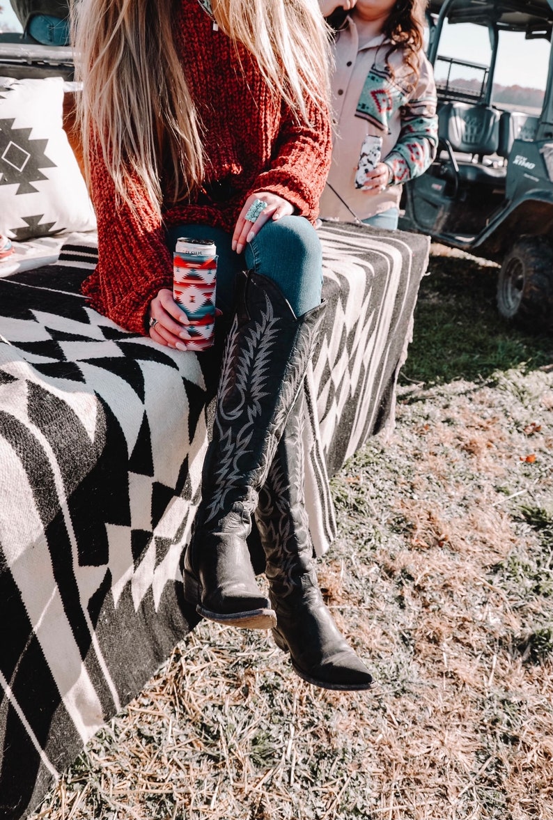 May include: A woman wearing a red sweater and blue jeans sits on a black and white patterned blanket. She is holding a can with a colorful patterned sleeve. She is wearing black cowboy boots with intricate designs.