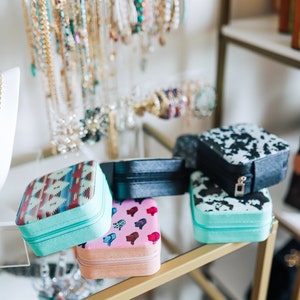 May include: Four small jewelry boxes with different patterns. The boxes are stacked on top of each other on a glass shelf with a gold metal frame. The boxes are turquoise, pink, black and white, and turquoise with a black and white cow print.