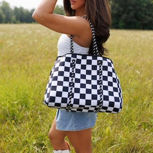 May include: A black and white checkered tote bag with black rope handles. The bag is carried by a person in a field of tall grass. The bag has a large capacity and a stylish design, suitable for various uses.