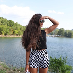 Pode incluir: Uma mulher vestindo um top preto sem mangas e shorts xadrez preto e branco está de pé perto de um lago. Ela está usando óculos de sol e tem cabelo castanho longo. Ela está segurando uma garrafa de água branca.