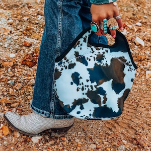 Puede incluir: Una bolsa de almuerzo aislada de vaca blanca y marrón con ribete negro. La bolsa tiene una cremallera y un asa. La bolsa la lleva una persona que lleva pantalones vaqueros azules y una camisa a rayas blancas y marrones.
