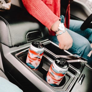 May include: Two cans of Celsius Sparkling Energy Drink in the cup holders of a car. The cans are wrapped in colorful patterned sleeves.