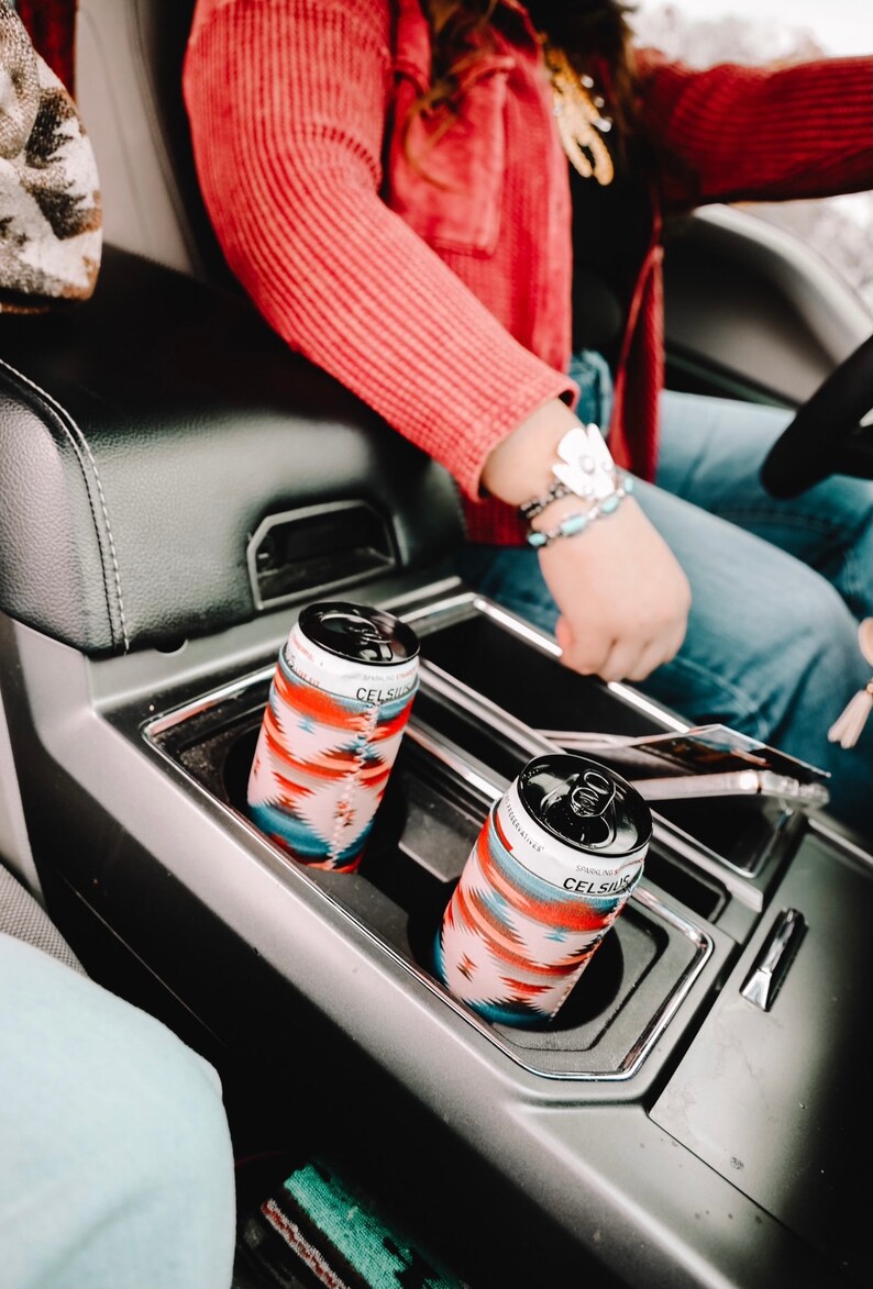 May include: Two cans of Celsius Sparkling Energy Drink in the cup holders of a car. The cans are wrapped in colorful patterned sleeves.