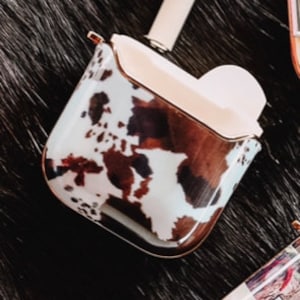 May include: A white and brown cow print phone case with a gold trim. The case is designed to fit wireless earbuds.