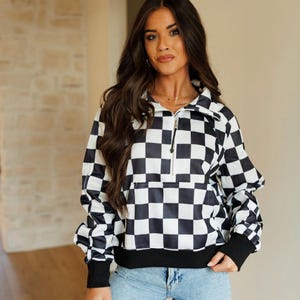 Puede incluir: Jersey con estampado de cuadros blancos y negros con cierre de media cremallera y bolsillo delantero. La parte superior de manga larga tiene puños negros y dobladillo negro. El modelo lleva vaqueros azul claro.