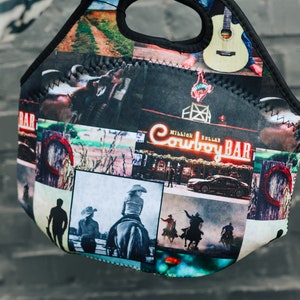 Peut inclure: Un sac à déjeuner en néoprène noir avec un collage coloré d'images, notamment des cowboys, des chevaux et une pancarte indiquant "Million Dollar Cowboy Bar".