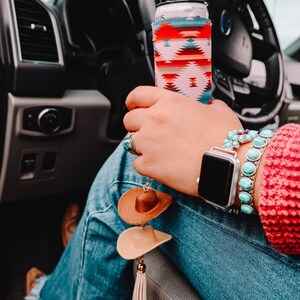 May include: A person wearing blue jeans and a red sweater with a turquoise and silver bracelet and a smartwatch. The person is holding a can with a colorful patterned can cooler. A brown and beige cowboy hat keychain hangs from the person's jeans.