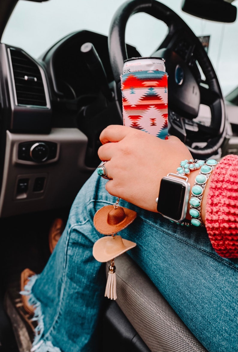 May include: A person wearing blue jeans and a red sweater with a turquoise and silver bracelet and a smartwatch. The person is holding a can with a colorful patterned can cooler. A brown and beige cowboy hat keychain hangs from the person's jeans.