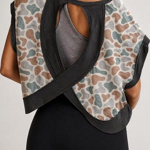 Könnte beinhalten: Ärmelloses Oberteil mit Camouflage-Muster in Braun-, Grün- und Beigetönen. Das Oberteil hat einen markanten Rückenausschnitt und schwarze Paspeln. Das Model trägt eine schwarze Leggings und einen grauen Sport-BH.