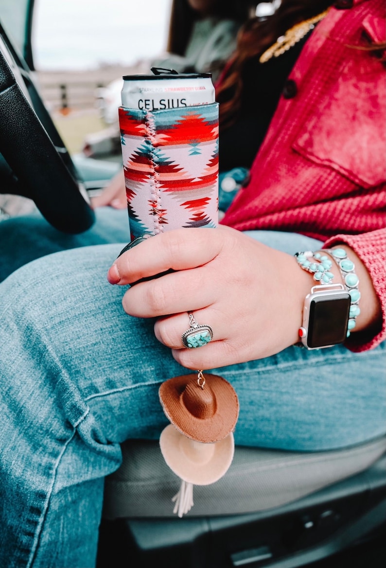 May include: A woman's hand holding a can of Celsius energy drink in a colorful patterned koozie. The can is partially visible and says "Celsius" on it. The woman is wearing a red sweater, blue jeans, and a turquoise and silver watch. A brown and beige cowboy hat keychain is hanging from her jeans.