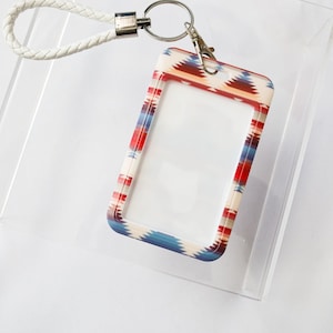 Puede incluir: Un cordón trenzado blanco con un llavero plateado y un portagafete con estampado colorido. El portagafete tiene una ventana de plástico transparente para insertar una tarjeta de identificación.
